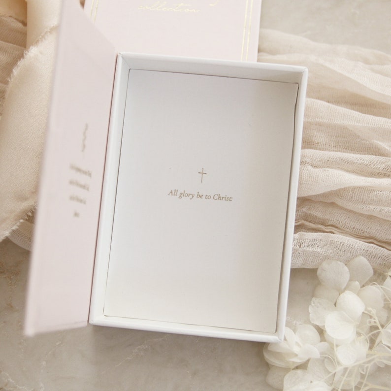 May include: A white box with a pink interior. The box is open and the inside is lined with white paper. The paper has a gold cross and the text "All glory be to Christ" printed on it.
