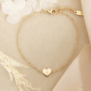 May include: A gold chain bracelet with a small heart charm featuring a cross design. The bracelet is on a light beige background.