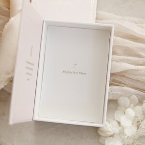 May include: A white box with a pink interior. The box is open and the inside is lined with white paper. The paper has a gold cross and the text "All glory be to Christ" printed on it.