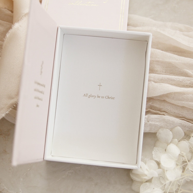 May include: A white box with a pink interior. The box is open and the inside is lined with white paper. The paper has a gold cross and the text "All glory be to Christ" printed on it.