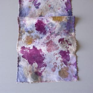 May include: Two pieces of fabric with a hand-dyed appearance. The fabric features a white base with splattered patterns in shades of purple, blue, and gold. The edges of the fabric are frayed, giving it a rustic look. The fabric is likely used for crafting or textile art.