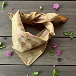 May include: A tan bandana with pink and gold speckles, knotted and displayed on a gray wooden surface. The bandana is surrounded by small purple flowers and green leaves, creating a natural aesthetic.