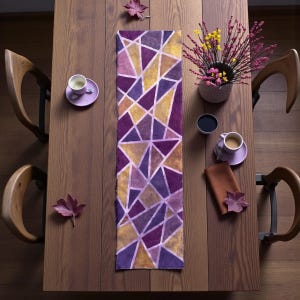 May include: Overhead view of a wooden dining table set for a meal. A purple and gold geometric patterned table runner is centered on the table. Two cups of coffee, a vase of flowers, and a napkin are also on the table.