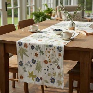 May include: A white fabric table runner featuring a botanical print of blue, orange, and green leaves and flowers. The runner is placed on a wooden table with two ceramic mugs, a small bowl of blueberries, a vase with greenery, and a book.