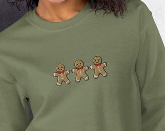 Pode incluir: Uma camisola verde com um design bordado de três homens de gengibre. Os homens de gengibre usam gravatas de laço vermelhas e têm detalhes de glacê branco.