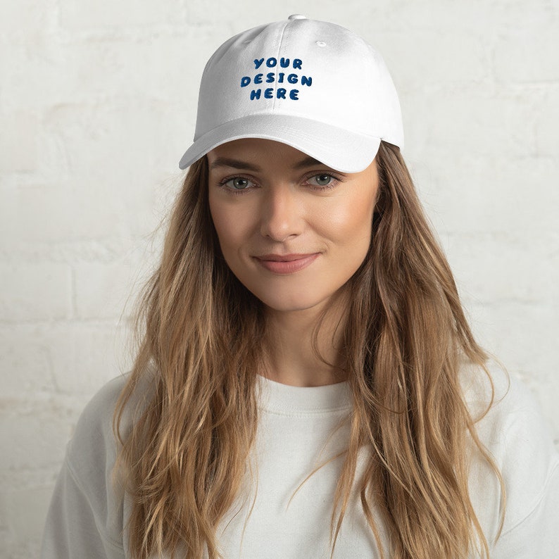 May include: A white baseball cap with a blue embroidered design that says "YOUR DESIGN HERE".