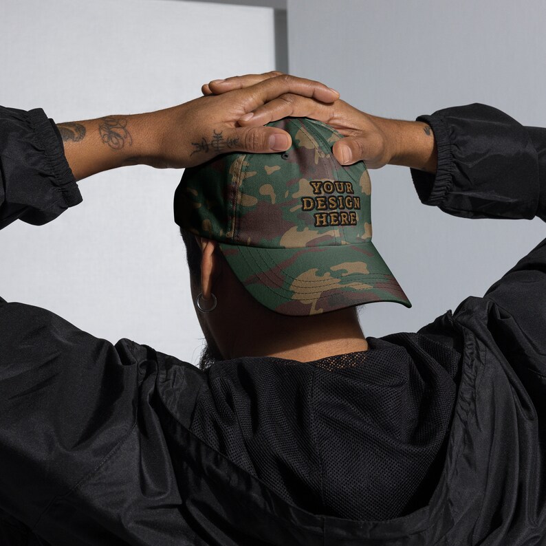May include: A camouflage baseball cap with the text "YOUR DESIGN HERE" embroidered in brown on the front.