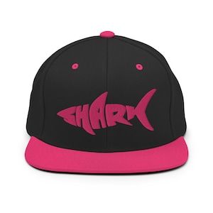 May include: A black and pink baseball cap with a pink shark silhouette and the word "SHARK" written inside the silhouette.