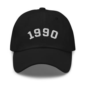 Puede incluir: Gorra de béisbol negra con el texto "1990" bordado en blanco en forma de arco.