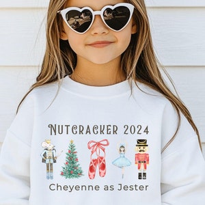 May include: White sweatshirt with a Nutcracker themed design. The design features a Christmas tree, a nutcracker, ballet shoes, and a ballerina. The text "Nutcracker 2024" and "Cheyenne as Jester" is printed on the sweatshirt.