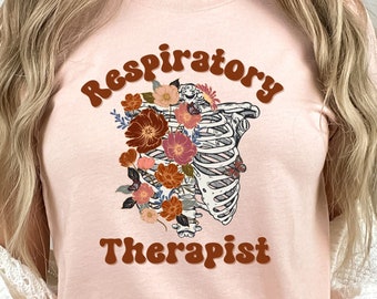 May include: A light pink t-shirt featuring a floral and anatomical design. The graphic includes a rib cage filled with colorful flowers and the words "Respiratory Therapist" in a retro font.