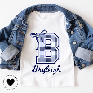 May include: White t-shirt with a navy blue gingham letter "B" design, a matching bow, and the name "Bryleigh" in script. A denim jacket is draped over the shirt. The shirt is from the "Charlie & KLO Girly Chic" shop.