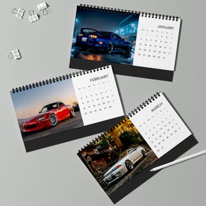 Puede incluir: Calendarios de escritorio con imágenes de coches deportivos. Los calendarios están encuadernados en espiral con bases negras e incluyen cuadrículas de fechas mensuales. Las imágenes muestran un coche azul por la noche, un coche rojo al atardecer y un coche blanco en una carretera.
