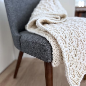 Puede incluir: Una manta de punto grueso de color crema sobre una silla gris tapizada. La manta tiene un diseño texturizado de punto de cable. La silla tiene patas de madera y un respaldo curvo, creando una escena acogedora y acogedora.