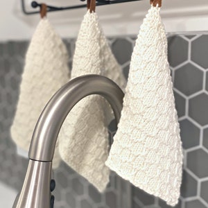 May include: Three cream-coloured, knitted washcloths hanging from a black metal bar with brown leather straps. The washcloths have a textured, patterned design. A stainless steel tap is in the foreground, with a grey hexagonal tile backsplash.
