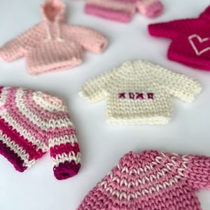 May include: A collection of miniature knitted sweaters in various shades of pink, white, and fuchsia. Some sweaters have striped patterns, a heart design, or the embroidered letters "XOXO". One sweater has a hood.