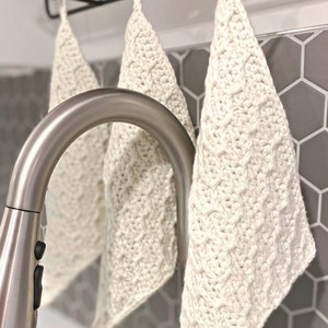 May include: Three cream-coloured, hand-crocheted washcloths hanging on a black rack. The washcloths feature a textured, cable-knit design. A stainless steel tap is in the foreground, with grey hexagonal tiles in the background.