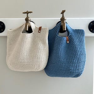 Può includere: Due borse tote all'uncinetto fatte a mano, una color crema e una blu, sono appese a ganci a forma di rubinetto in ottone su una tavola di legno bianca. Le borse hanno dettagli in pelle marrone e un aspetto robusto e strutturato.