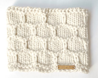 May include: A cream-coloured, chunky knit cowl featuring a cable stitch design. The thick, soft yarn creates a textured surface. A small, rectangular wooden tag with the text "(OBC) est. 2018" is sewn on the bottom right corner.