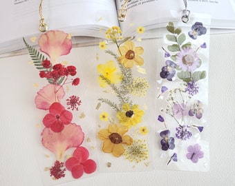 May include: Three clear bookmarks with pressed flowers and gold accents. The bookmarks feature a variety of flowers in shades of pink, yellow, and purple.