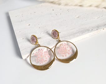 Könnte beinhalten: Ein Paar goldene Creolen mit gepressten rosa Blüten, die in klarem Harz eingeschlossen sind. Die Ohrringe haben ein zartes, feminines Design.