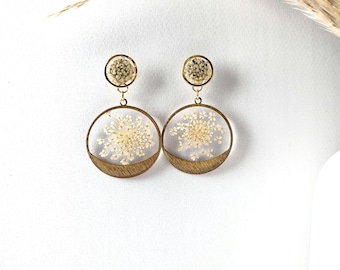 Puede incluir: Un par de pendientes de tono dorado con un diseño redondo de resina transparente que presenta delicadas flores blancas. Los pendientes tienen un acento de madera en forma de semicírculo.