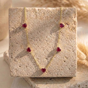 May include: A gold chain necklace featuring five evenly spaced, small, heart-shaped red gemstones. The delicate metal chain connects each vibrant crimson heart, creating a symmetrical design that hangs in a gentle V-shape across the surface.
