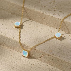 May include: A delicate gold necklace featuring three square opal pendants. The opals have a subtle iridescent quality, set in gold bezels. The necklace chain is fine and elegant, perfect for layering or wearing alone. The jewelry is displayed on a textured, neutral surface.