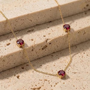 May include: A delicate gold necklace featuring three oval-cut, deep red gemstones. The gemstones are evenly spaced along the fine gold chain. The necklace is displayed on a textured, light beige surface, highlighting the jewelry's elegant design.