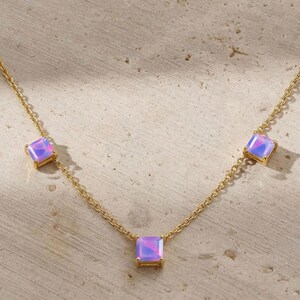 May include: A delicate gold necklace featuring three square, iridescent, lavender-colored gemstones. The gemstones are set in gold bezels and spaced evenly along the gold chain. The necklace is displayed on a neutral, textured surface.