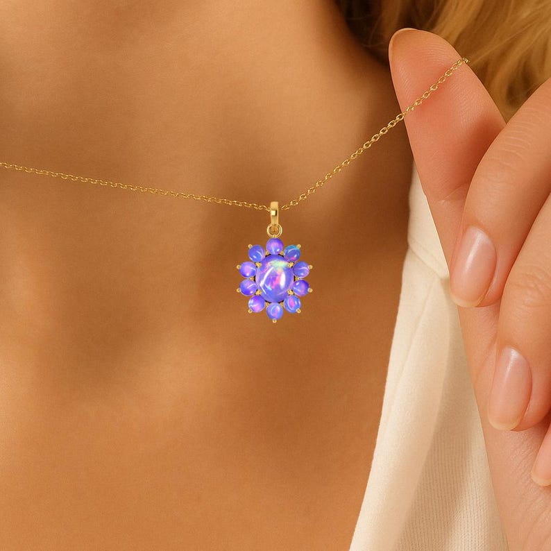 May include: A gold-colored necklace featuring a pendant with a flower-like design. The pendant is composed of a central oval-shaped, light purple stone surrounded by smaller, similarly colored stones. The necklace is displayed against a neutral background.