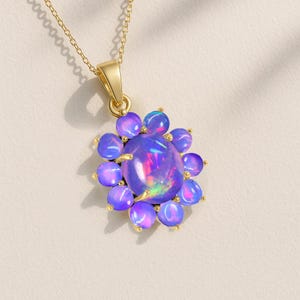 May include: A gold-colored necklace featuring a pendant with a large, iridescent opal stone at the center, surrounded by smaller, round, blue-purple stones. The pendant is suspended from a delicate gold chain.