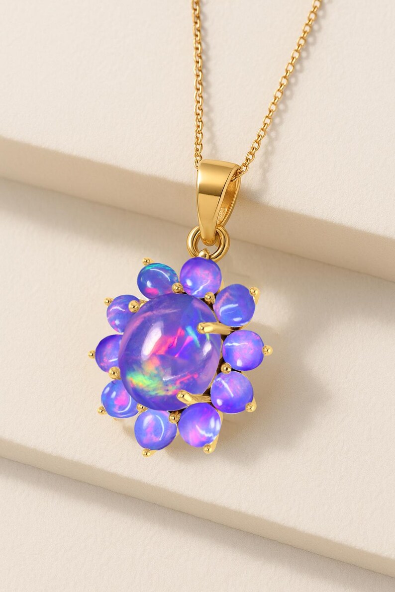 May include: A gold-colored necklace featuring a pendant with a large, oval, iridescent purple gemstone surrounded by smaller, round, purple gemstones. The pendant is suspended from a delicate gold chain.
