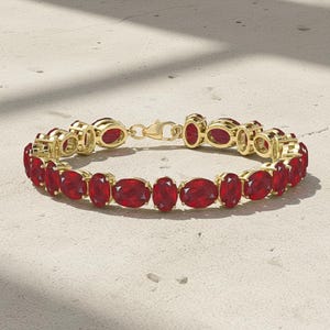 May include: A gold bracelet with oval-cut, deep red gemstones. The bracelet is secured with a gold clasp and is shown on a light surface. The gemstones are evenly spaced and set in gold bezels.