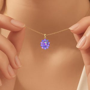 May include: A gold necklace with a pendant featuring a cluster of purple opals. The pendant is in the shape of a flower, with a central opal surrounded by smaller opals. The necklace is held by a person.
