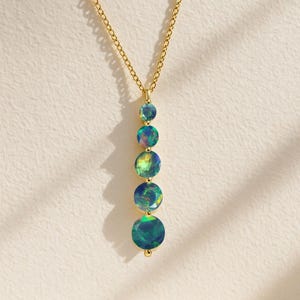 Puede incluir: Un collar dorado con una serie de piedras preciosas redondas e iridiscentes en tonos azules, verdes y amarillos. Las piedras están dispuestas en línea vertical, suspendidas de una delicada cadena. El collar se muestra sobre un fondo neutro.