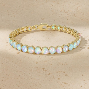 May include: A gold bracelet with oval opals. The opals display a mix of colours, including white, blue, and pink. The bracelet has a clasp closure and is shown on a light-coloured surface.