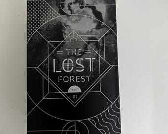 Op de afbeelding: Een zwart tarotkaartspel met de titel "THE LOST FOREST" in witte, gestileerde letters. Het ontwerp van de omslag omvat geometrische vormen en het woord "TAROT". De achtergrond heeft een wolkig, atmosferisch ontwerp en creëert een mystieke uitstraling.