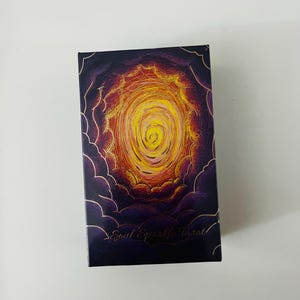 Könnte beinhalten: Rechteckige Tarotkartenbox mit dunkelviolettem Hintergrund und einem zentralen Design eines wirbelnden gelb-orangen Wirbels. Die Box ist von einem wolkenartigen Rand umgeben, und der Text "Soul Sparkle Tarot" ist in Gold gedruckt.
