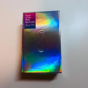 May include: A holographic tarot card box with the text "Tarot of the Holy Spectrum Infinity Edition" on a pink label. The box has a raised infinity symbol and reflects a rainbow of colours.