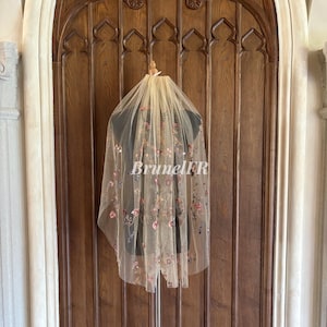 Puede incluir: Un velo de tul marfil delicado con bordados florales en tonos rosa, azul y verde. El velo se muestra en un maniquí sobre un fondo de madera.