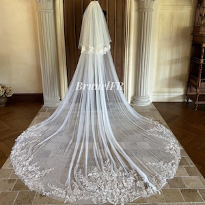 Peut inclure: Un long voile de mariée blanc avec une délicate bordure en dentelle florale. Le voile est présenté sur un mannequin devant une porte en bois. La traîne du voile s'étend sur le sol, mettant en valeur le design en dentelle.