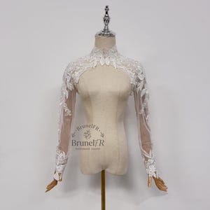 Peut inclure: Boléro de mariée en dentelle blanche à manches longues, présenté sur un mannequin. Le boléro présente des motifs floraux et de feuilles complexes, avec des détails délicats le long de l'encolure et des poignets. Le logo Brunel FR est visible.