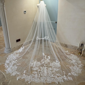 Può includere: Un velo da sposa bianco con un bordo in pizzo floreale. Il velo è lungo e fluido, e il pizzo è delicato e intricato.