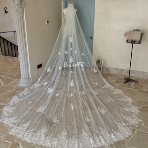 Peut inclure: Un voile de mariée blanc avec une délicate broderie de dentelle florale. Le voile est long et fluide, avec un bord festonné. Le voile est présenté sur un mannequin dans une pièce avec une porte bleu clair.