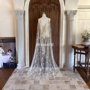 Può includere: Un velo da sposa bianco e trasparente con ricami dorati di stelle e lune. Il velo è drappeggiato su un manichino in una stanza con una porta di legno e colonne.