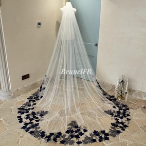 Op de afbeelding: Een lange witte bruids sluier met een marineblauwe en gouden florale kantversiering. De sluier wordt getoond op een mannequin in een kamer met een betegelde vloer.