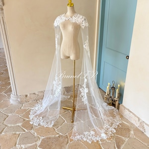 Peut inclure: Une cape de mariée blanche avec des détails de dentelle florale. La cape présente un col montant et de longues manches fluides, présentée sur un mannequin. Le design comprend des motifs délicats sur les bords et les manches, créant un look élégant et romantique.
