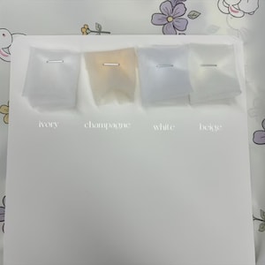 May include: A display of four fabric swatches in ivory, champagne, white, and beige, each secured with a silver staple. The colors are labeled below each swatch against a white background. The background has a floral pattern.