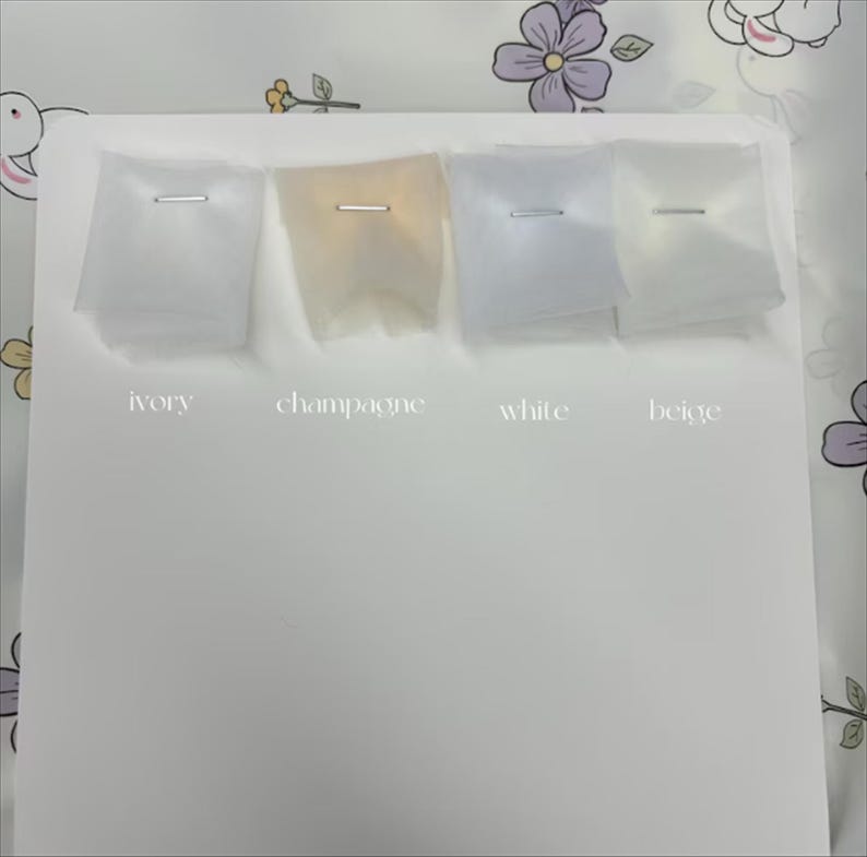 May include: A display of four fabric swatches in ivory, champagne, white, and beige, each secured with a silver staple. The colors are labeled below each swatch against a white background. The background has a floral pattern.